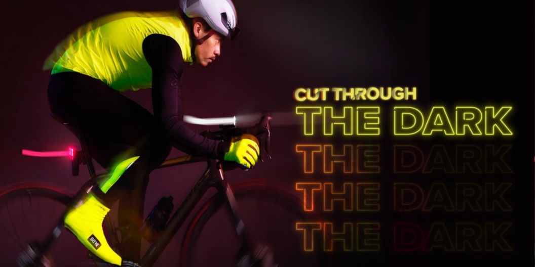 A rider in the dark with high-vis cycling apparel and front and rear lights pedals toward a graphic neon light display showing the phrase “cut through the dark.”